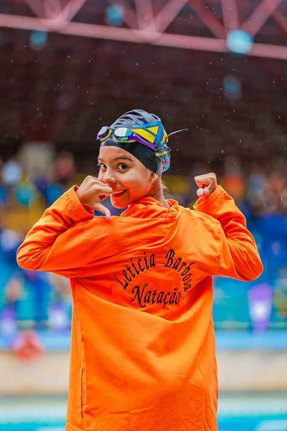 Lelê de roupão laranja com o nome Letícia Barbosa bordado, fazendo pose na beira da piscina